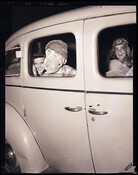Children in costume, dressed for Halloween, sitting in the back of a car. The child closest to the rear window wears a mask. The other two children smile at the camera from inside a car. The photograph was taken in the dark, likely night, with a bright flash. Photograph by Robert F. Kniesche (1906-1976).