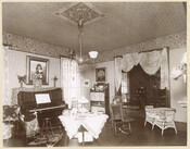 View of a sitting room in La Paix, an estate located on York Road near Sheppard Station in Towson, Maryland. The room features a piano as well as a variety of furniture and visible in the background is an entrance to a dining room. Designed by Baltimore architect John Appleton Wilson for the Lawrence Turnbull…