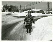 View of a man wearing a plaid coat and holding a paper bag as he walks through the snow. A 7-Eleven convenience store is pictured directly behind him.