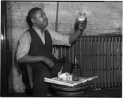 An unidentified man seated and holding a National Bohemian (Natty Boh) beer in a mason jar glass. The beer bottle sits atop a newspaper on a small table.