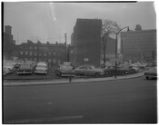 View of a parking lot full of cars at the intersection of South Paca Street and Camden Street in Baltimore, Maryland. Shows row houses and warehouse in background.