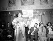 Mahalia Jackson singing into a microphone at a church, possibly Gillis Memorial Church in Baltimore, Maryland, as Gillis's pastor Reverend Theodore Jackson appears in a related photograph. Men and women are seated behind Mahalia Jackson, clapping and singing. A sign above her head reads, "Have you prayed about it? Prayer changes things."