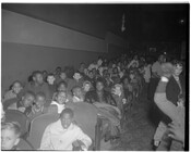 People sitting in rows in an unidentified theater. The crowd is mostly children, but several adults are also present.