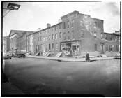 Street view of North Arlington Avenue, Baltimore, Maryland. A grocery store is located on the corner and a pedestrian is crossing the street. Several people are seen standing inside the doorways of the buildings.