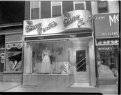 Street view of The Charm Centre, a clothing and accessories store located at 1811 Pennsylvania Avenue in Baltimore, Maryland. The store was owned by William Lloyd "Little Willie" Adams (1914-2011), a prominent Black businessman, and his wife Victorine Q. Adams. The store fronts of Fredericks (shoes) and Morgan's (eye examiners) are also visible.