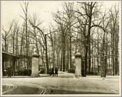 Entrance to Druid Hill Park at Druid Hill Avenue and Fulton Street, Baltimore, Maryland. Shows two stone pillars, ironwork archways, streetcar tracks, a "Drive Slow" sign, and three men, two of whom are walking into the park.