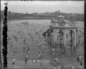 A view of bathers at the Clifton Park swimming pool in Baltimore, Maryland. A structure that has two staircases leading to a platform with sliding boards is visible in the right foreground. Established in 1894, Clifton Park is a public park located in the northeastern region of Baltimore City, Maryland.
